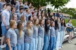 Group of high school bleachers wearing matching gray t-shirts stand on outdoor bleachers