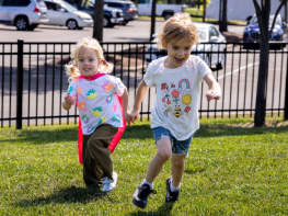 Two young children, one wearing a cape, run across a grassy field
