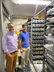 Young man in blue polo and middle aged man in purple shirt stand in storage room with long, round, metal tubes
