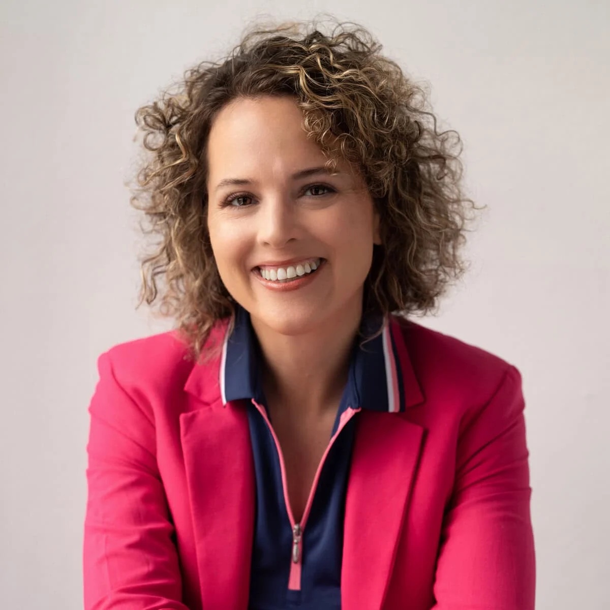 A smiling woman with curly hear in a pink blazer poses in front of a beige background for a professional headshot