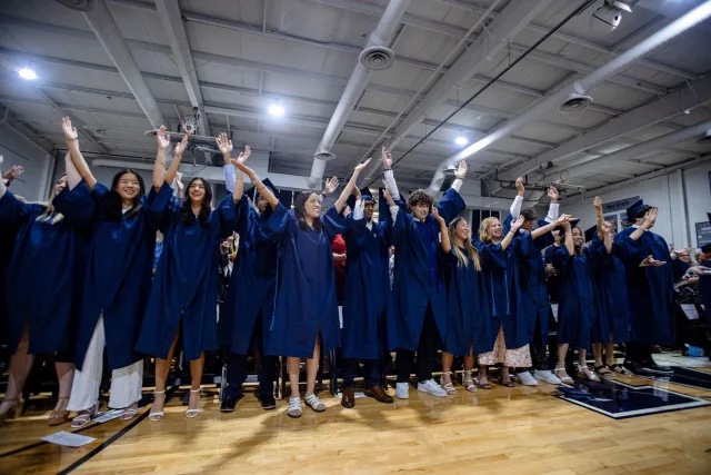High school seniors in deep blue graduation robes reach their hands toward the ceiling