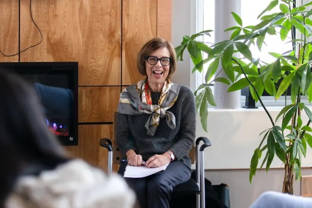 A woman smiles while seated in a chair in the front of a room