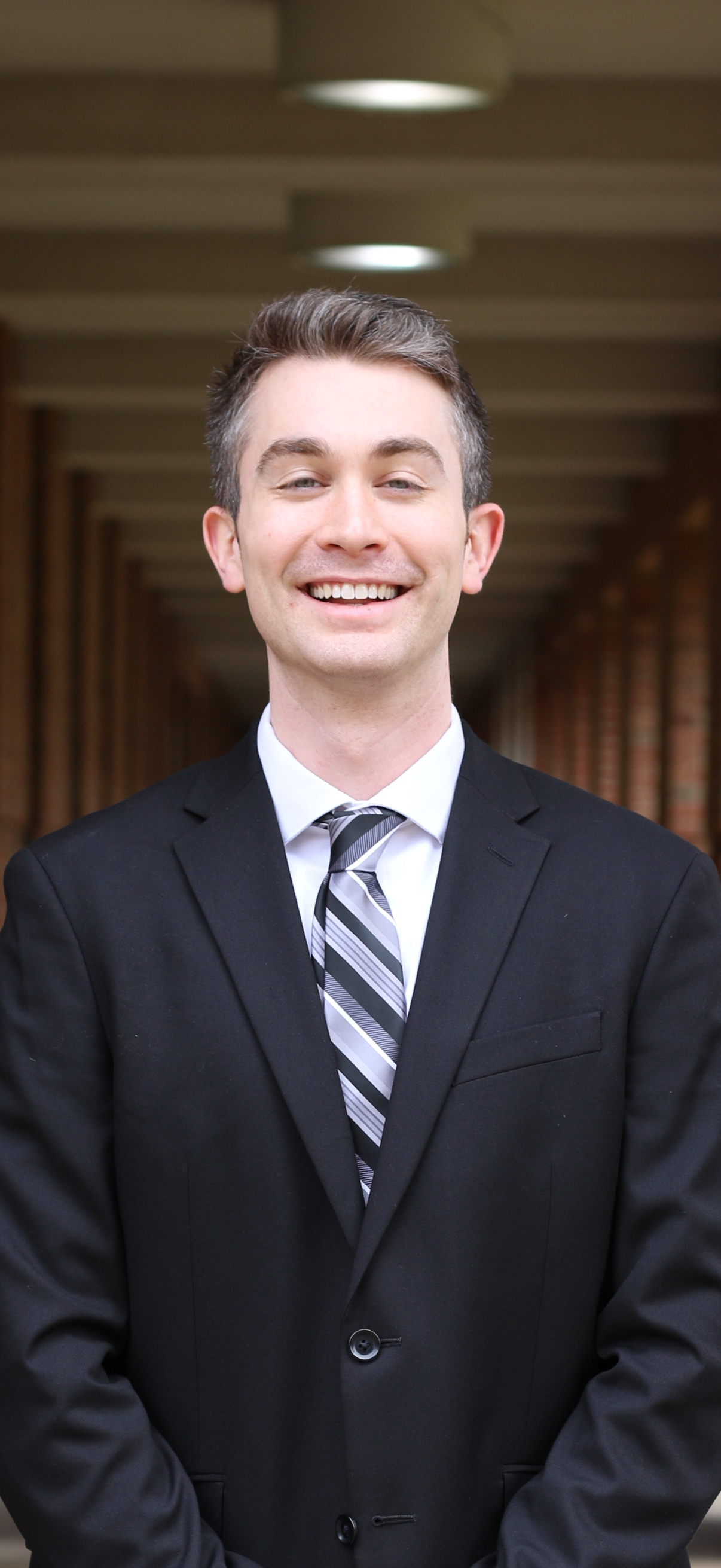 A smiling man in a suit or tie poses for a headshot in an architectural hallway